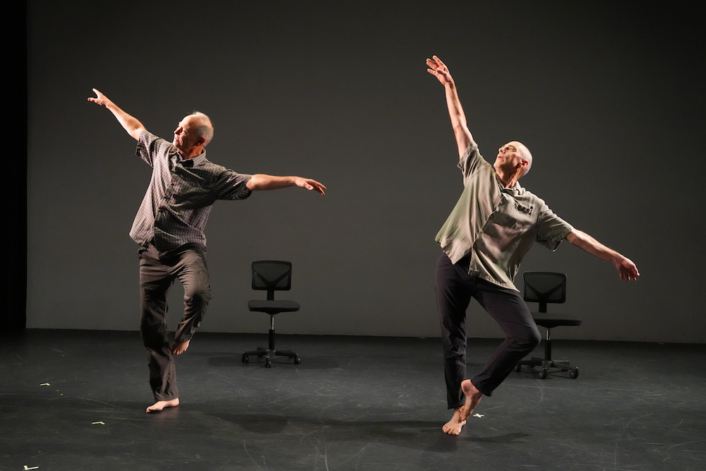 two older men with light skin wearing short sleeved light shirts and dark grey pants dance. They stand on one leg, leaning away from it, as the other leg is lifted with it's foot touching the standing legs knee. There are two chairs behind the duo.